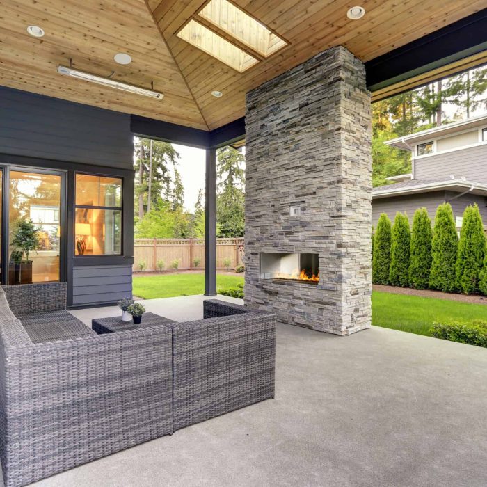 New modern home features a backyard with covered patio accented with stone fireplace, vaulted ceiling with skylights and furnished with gray wicker sofa placed on concrete floor. Northwest, USA
